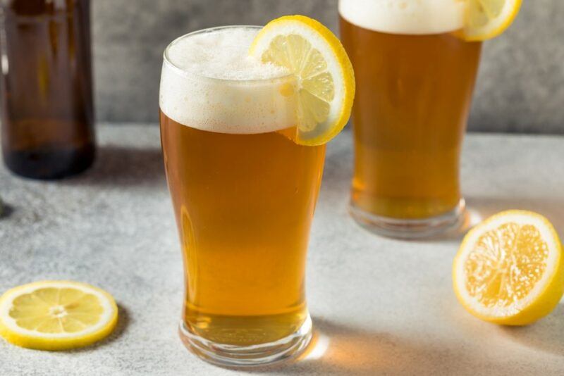 Two pint glasses containing shandies, each garnished with a lemon slice, with more lemons on the table