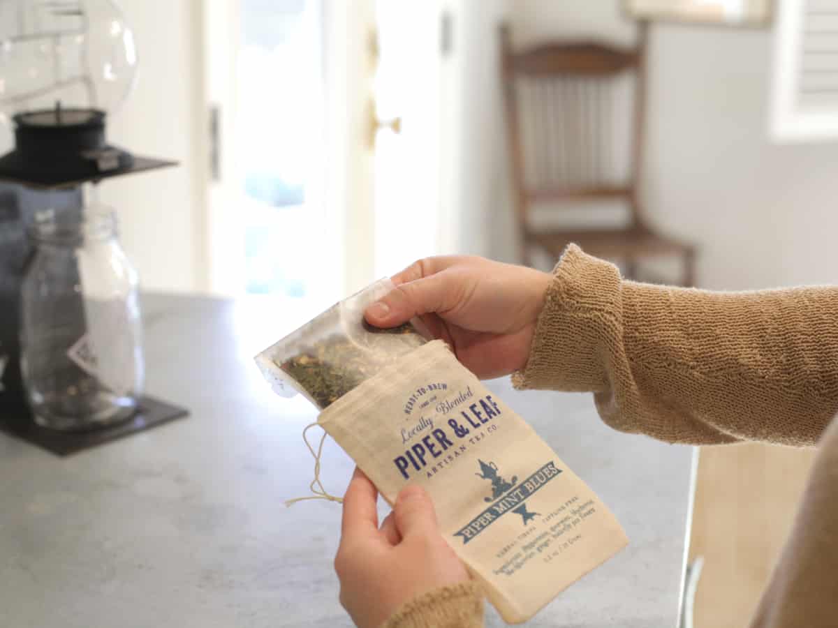 Person's hands and forearms on the right side of the photo holding pouch of tea with bag that states Piper and leaf, Piper Mint Blues. Background is a kitchen counter 