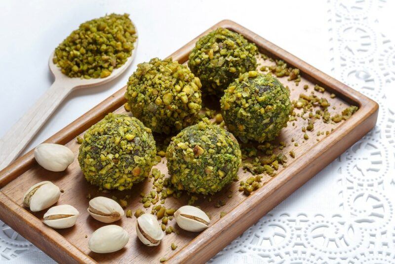 A wooden board with edges containing five pistacho truffles and some pistachio nuts, next to a spoon of ground pistachios