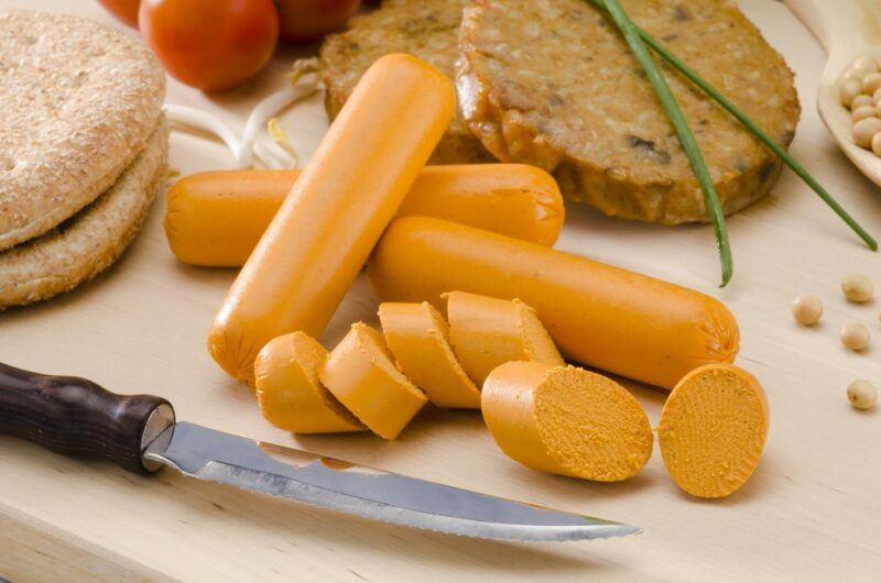 A small pile of plant-based sausages, one that has been cut in to pieces, in front of plant-based burger patties