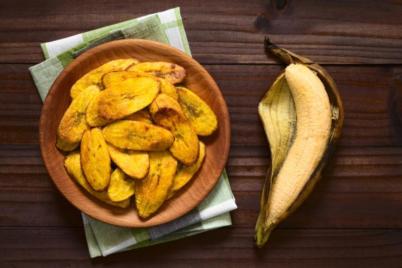 Roasted plantains in a wooden bowl with a fresh plantain on a table