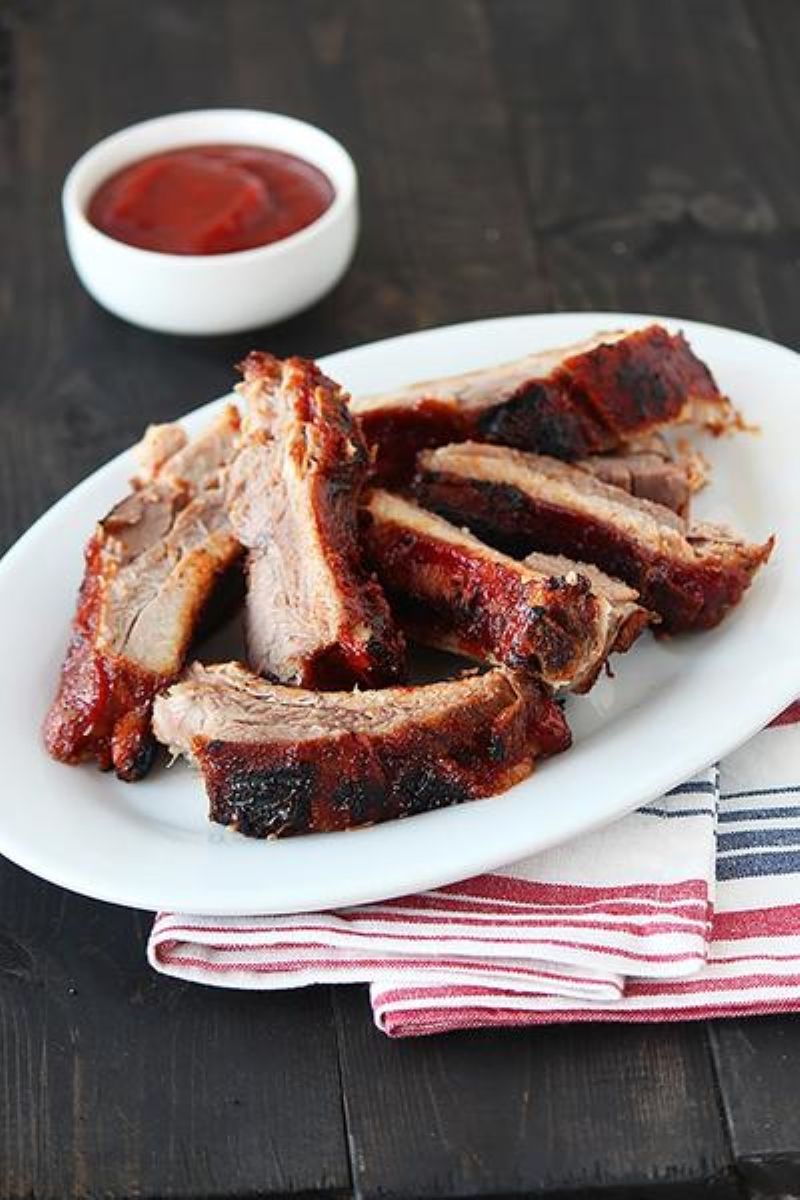 White oval plate of ribs that have been cut up into individual bones.  Below the plate is a red, blue, and white napkin. In the background is a small bowl of sauce out of focus.