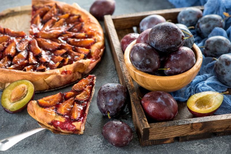 A plum pie with one piece cut out of it, next to a dish of plums and a bowl of them