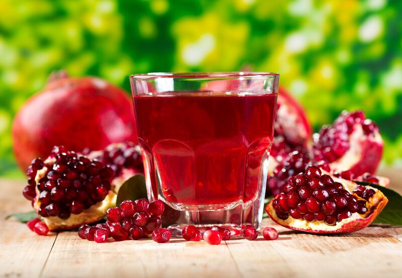 This photo shows a glass of pomegranate juice near some open pomegranates and a whole pomegranate in the background on a wooden table.