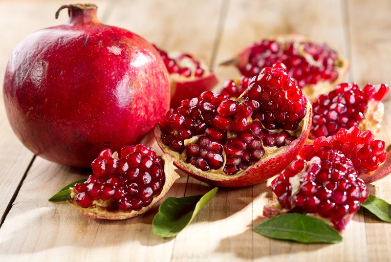 An open pomegranate fruit lies on a wooden countertop next to an unopened pomegranate fruit and some green leaves.