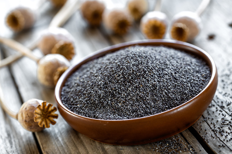 Brown ceramic bowl full of poppy seeds on a wooden table with loose poppy seeds and poppy seed pods around it.