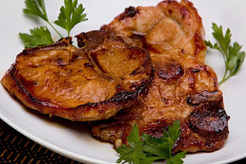 Two brown cooked pork chops rest on a white ceramic plate with three sprigs of parsley.