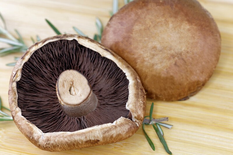 This photo shows an upright and an overturned Portobello mushroom on a wooden table near some sprigs of rosemary.