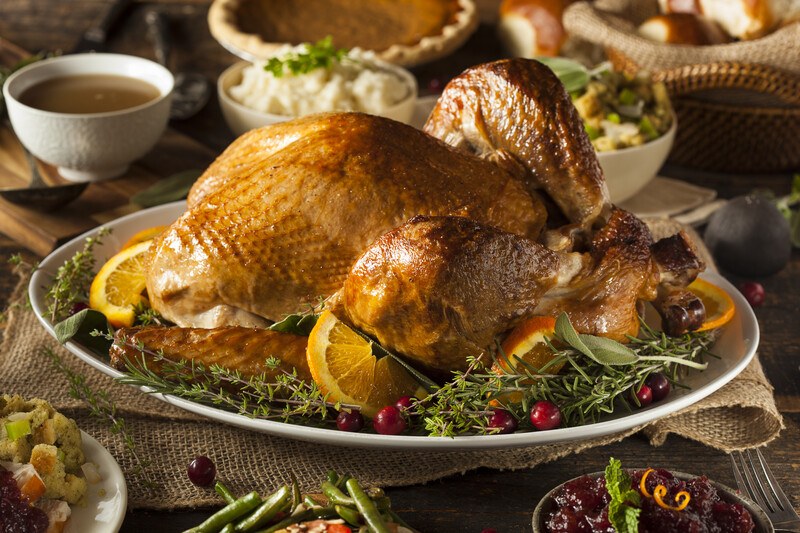 This photo shows a cooked, browned turkey on a platter with rosemary, cranberries, and orange slices resting on a piece of brown burlap cloth in the middle of a wooden dining table, surrounded by rolls, a pumpkin pie, and other traditional Thanksgiving dishes.