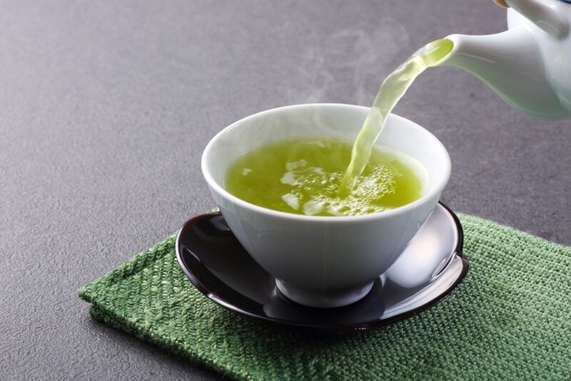 A stream of green tea pours from a white tea kettle into a white cup in a black saucer, on a green cloth, on a gray surface.