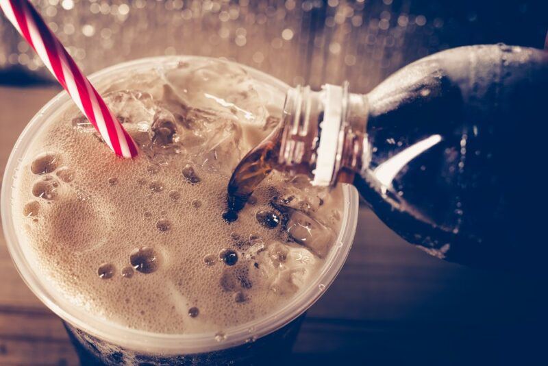 Coke being poured from a bottle into a plastic glass, complete with ice and a red and white straw