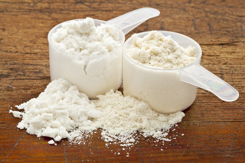 Two clear plastic scoops are filled with overflowing protein powder on a wooden countertop.