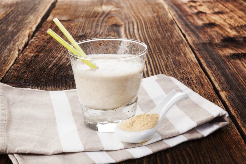 A white protein shake in a glass next to a spoon of protein powder