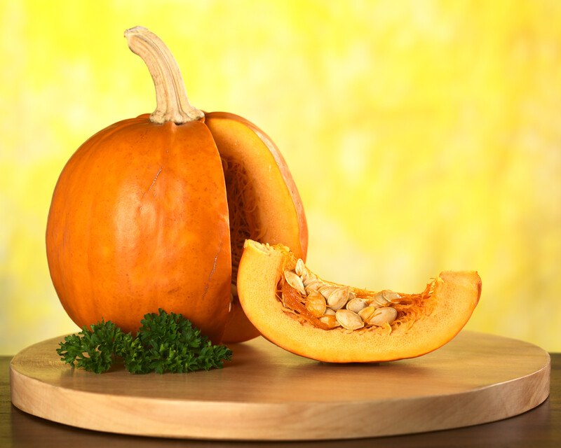 An orange pumpkin with a wedge cut from it rests on a wooden disk with some green leafy garnish against a yellow background.