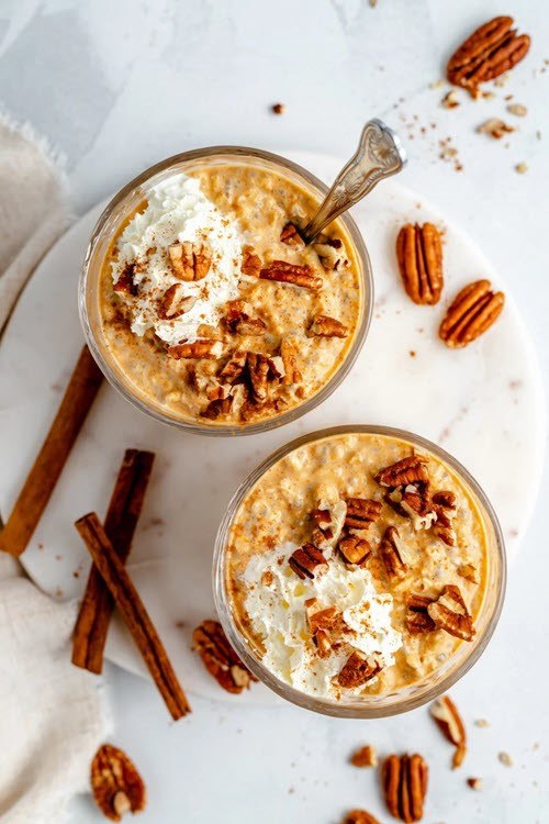 Two containers of pumpkin pie overnight oats, one with a spoon, next to cinnamon sticks and pecans