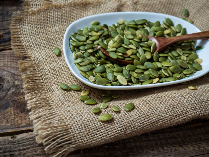 A white dish filled with green shelled pumpkin seeds and a wooden spoon rests on a piece of tan burlap cloth on a wooden surface.