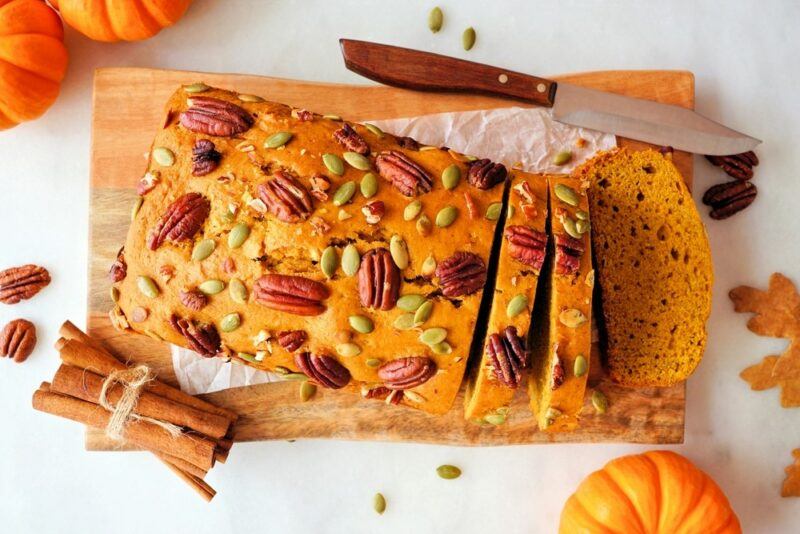 A white table with a sliced loaf of pumpkin bread that has been topped with nuts and seeds