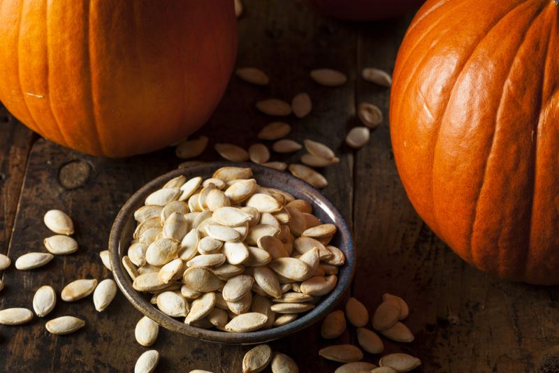 A bowl of unroasted pumpkin seeds, with pumpkin seeds scattered on the table and two orange pumpkins