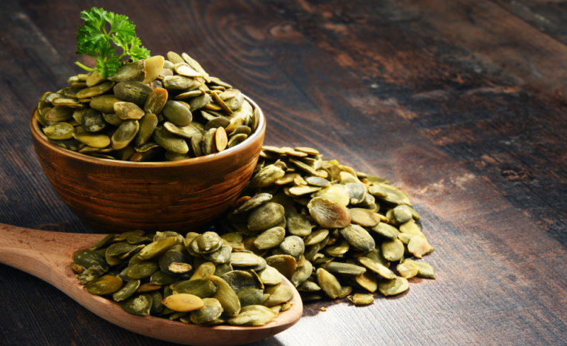 A wooden table with a bowl of pumpkin seeds, a wooden spoon of the seeds and more seeds on the table