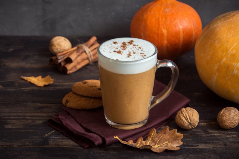 A glass of pumpkin spice latte next to pumpkins, fall leaves, walnuts and cinnamon