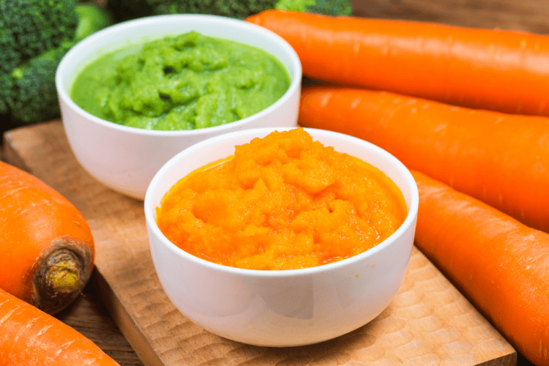 A white bowl of pureed carrots and another of pureed broccoli, next to many carrots and a small amount of broccoli