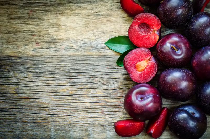 A selection of bright purple plums on the right hand side of a wooden board. Some have been cut open to reveal red flesh