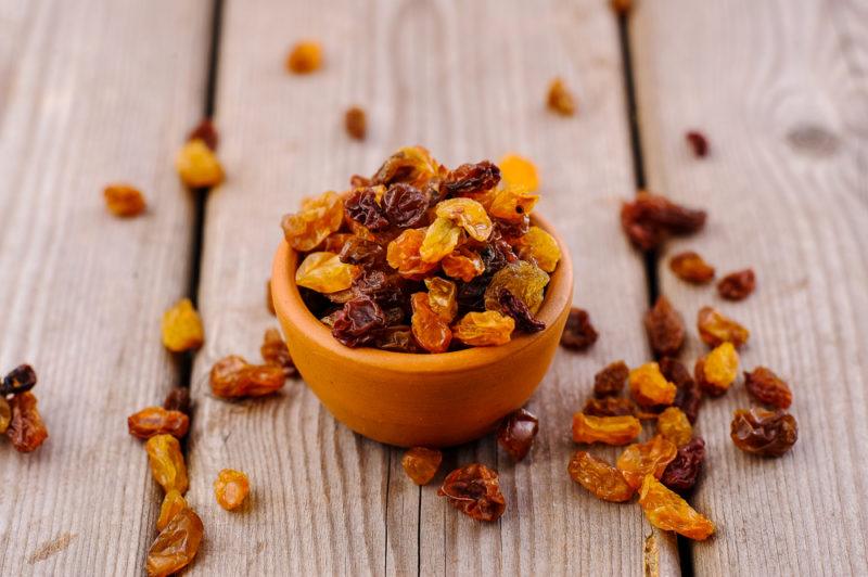 A pottery bowl containing raisins on a wooden table with raisins scatered around