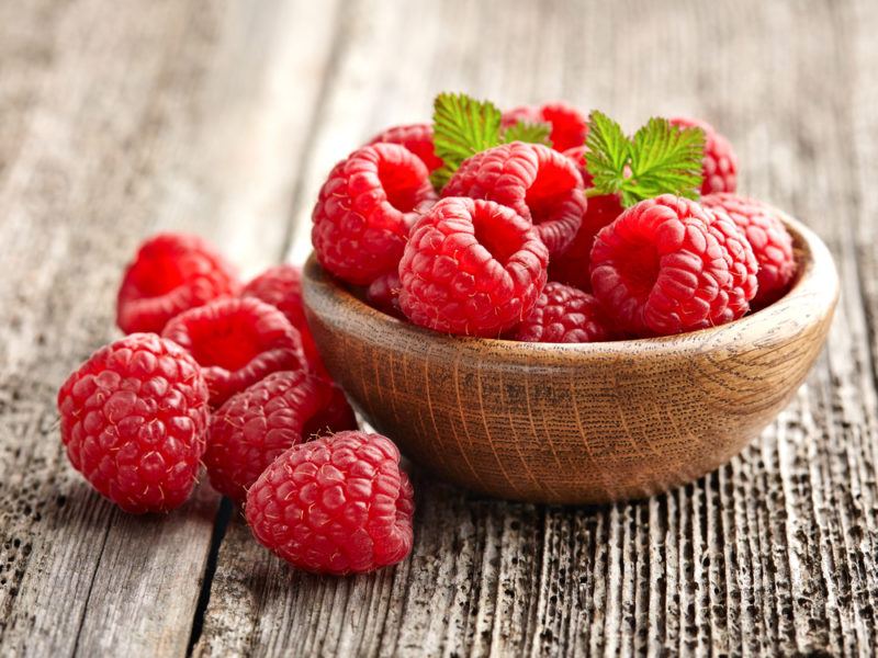 A wooden bowl of raspberries on a wooden table with a few berries on the table itself