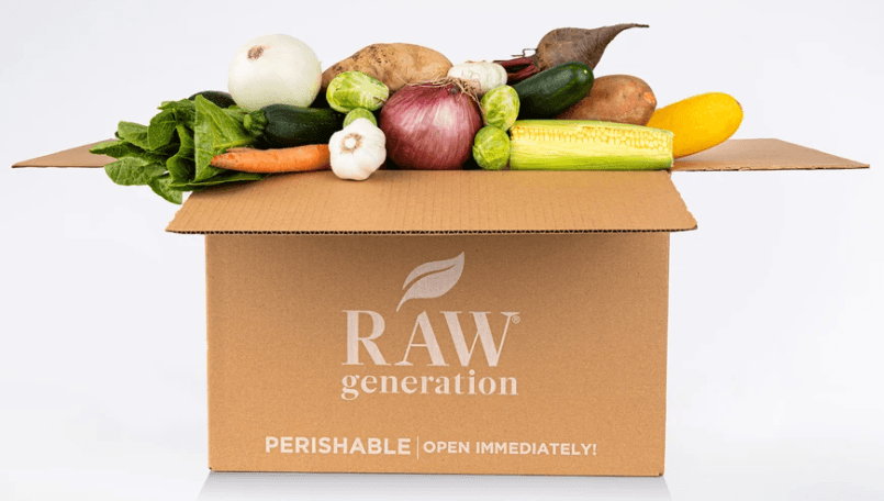 CARDBOARD BOX WITH RAW GENERATION STAMPED ON THE OUTSIDE OF THE BOX, THE BOX IS OVERFLOWING WITH VEGETABLES LIKE ONIONS, BEETS, BRUSSELS SPROUTS, CORN, AND GARLIC 