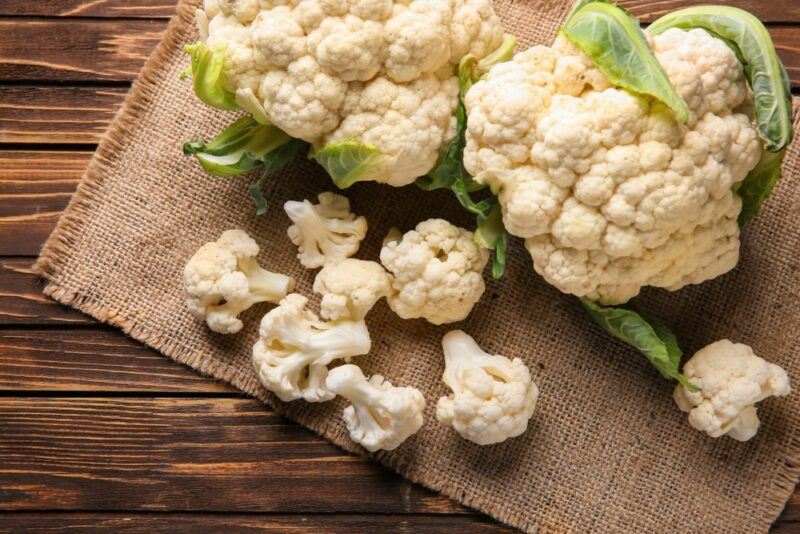 A wooden table with a woven cloth, two large heads of cauliflower and small pieces of cauliflower