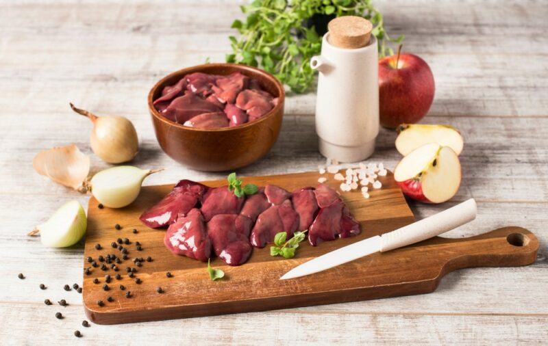 A wooden tray with pieces of chicken liver, in front of a bowl of chicken liver, with various ingredients scattered around, including herbs, onions, and black pepper