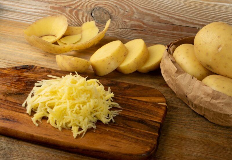 A wooden board with shredded raw potato, in front of potato slices and a bowl of potatoes