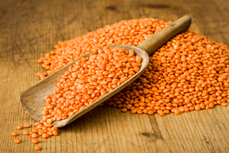 A wooden table with a large pile of red lentils and a scoop of the lentils