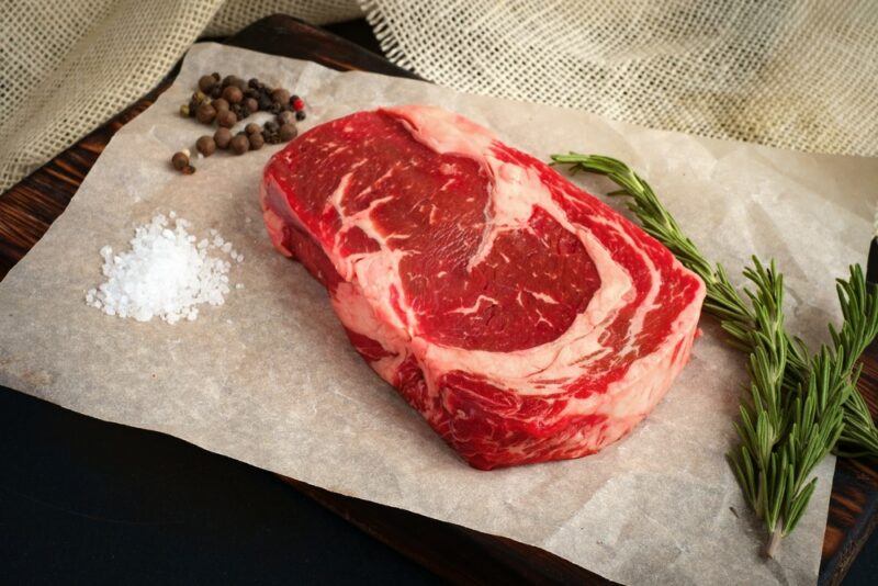 A piece of cooking paper that contains a raw ribeye steak next to herbs, salt, and pepper