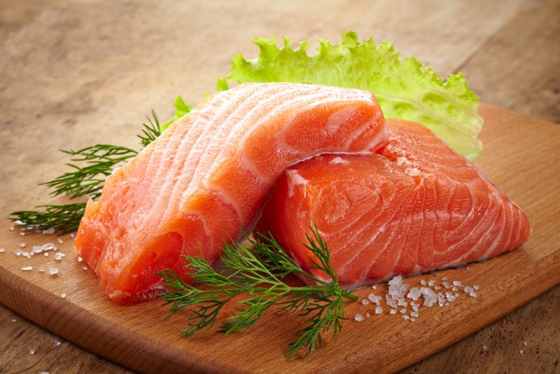 Two pieces of raw salmon on a cutting board with greenery and salt