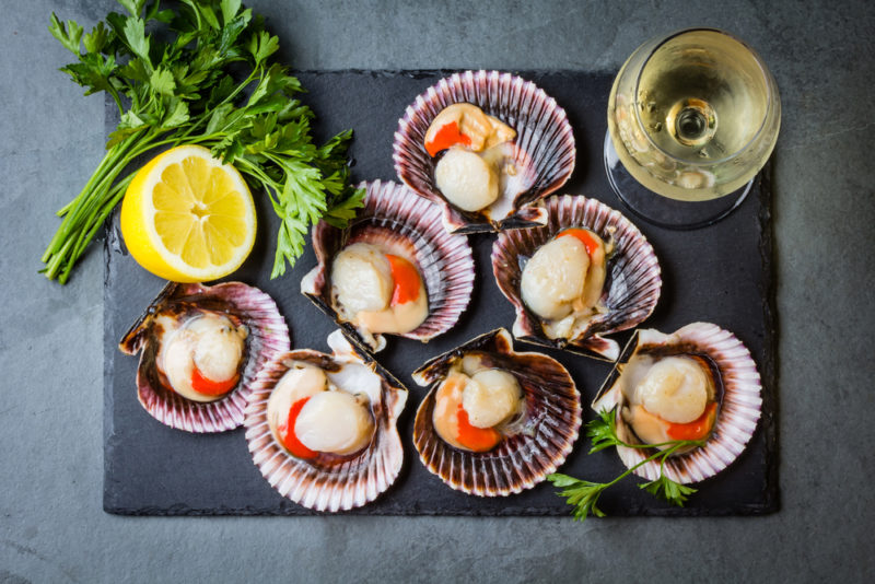 A selection of raw scallops in their shells on a black slate with parsley, lemons, and a glass of wine