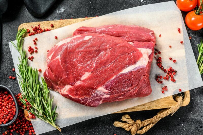 A white plate on a bag that contains a raw piece of top round steak next to rosemary and red peppercorns