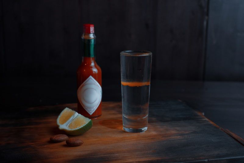 A cocktail glass containing a red dog shot, which includes a red layer of Tabasco sauce, in front of a Tabasco bottle and a wedge of lime