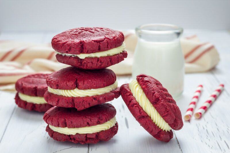 A pile of three red velvet cookies with cream cheese stuffing, plus another couple nearby and a small glass container of milk