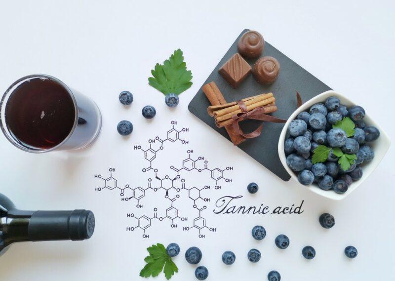 A glass of red wine, a bottle of red wine, some chocolate, and some blueberries, surrounding the chemical structure for tannic acid