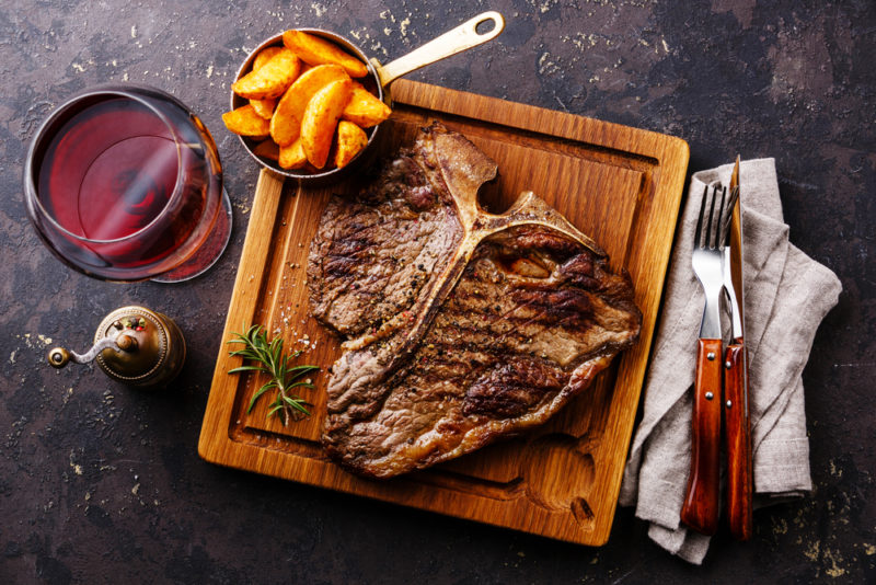 A square wooden board with a t-bone steak and fries, next to a knife, fork and glass of red wine