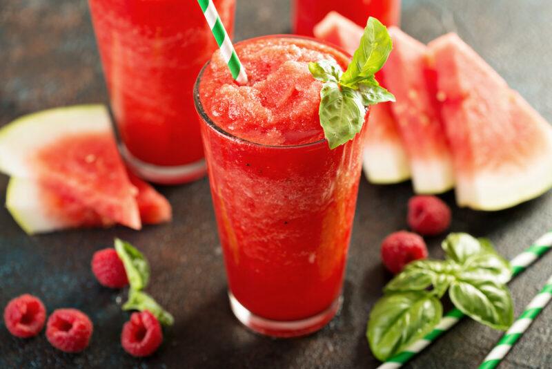 A cold summer drink made using watermelon and basil on a table next to some watermelon