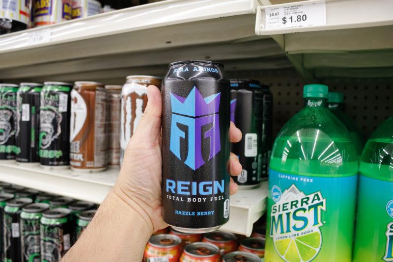 Someone holding a can of Reign Total Body Fuel in front of a grocery store shelf with other types of soda and energy drink