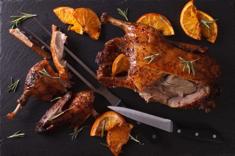 Roast duck on a black background that has been carved. There is a knife, a carving fork, and various oranges on the plate too.