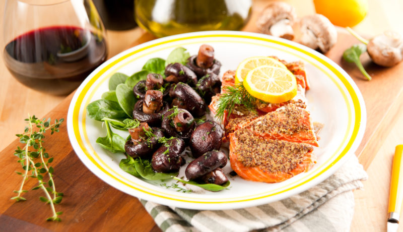 A white plate with yellow around the rim, containing roast salmon and cooked mushrooms, next to a stemless glass of pinot noir