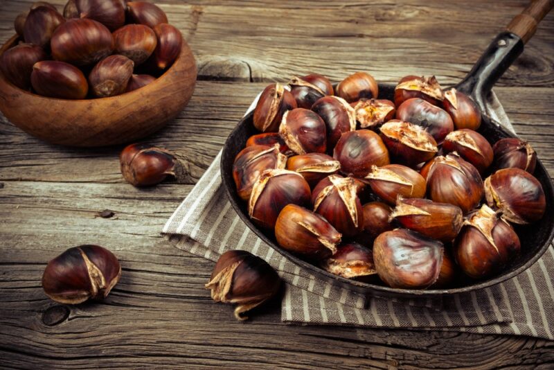 A large flat dish of roasted chestnuts in a frypan, next to a small wooden bowl of uncooked ones