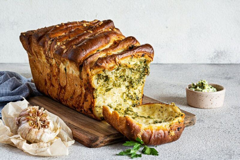 A large loaf containing rosemary and garlic pull apart bread on a table next to garlic and a small container of rosemary butter