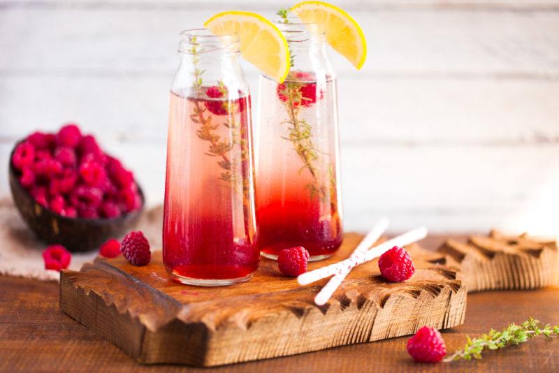 Two tall glasses containing rosemary raspberry lemonade with the ingredients scattered on the table