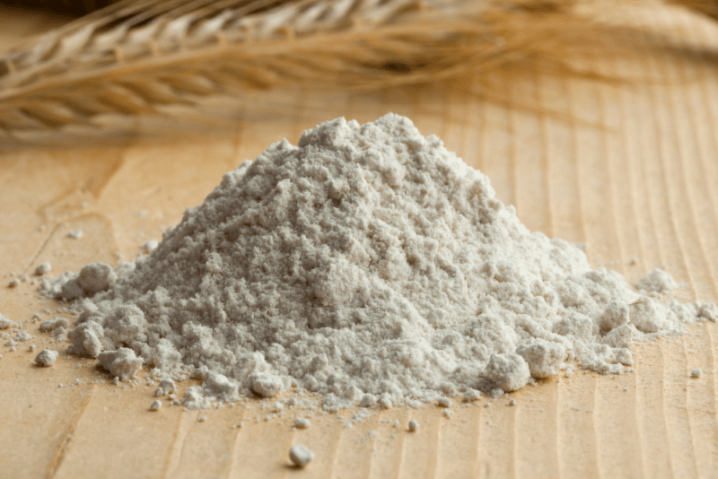 A wooden table with a pile of rye flour and a rye stalk in the background.