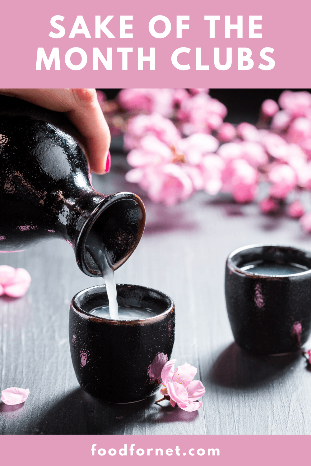 A hand pouring sake from a black jug into small black cups with pink blossoms in the background.